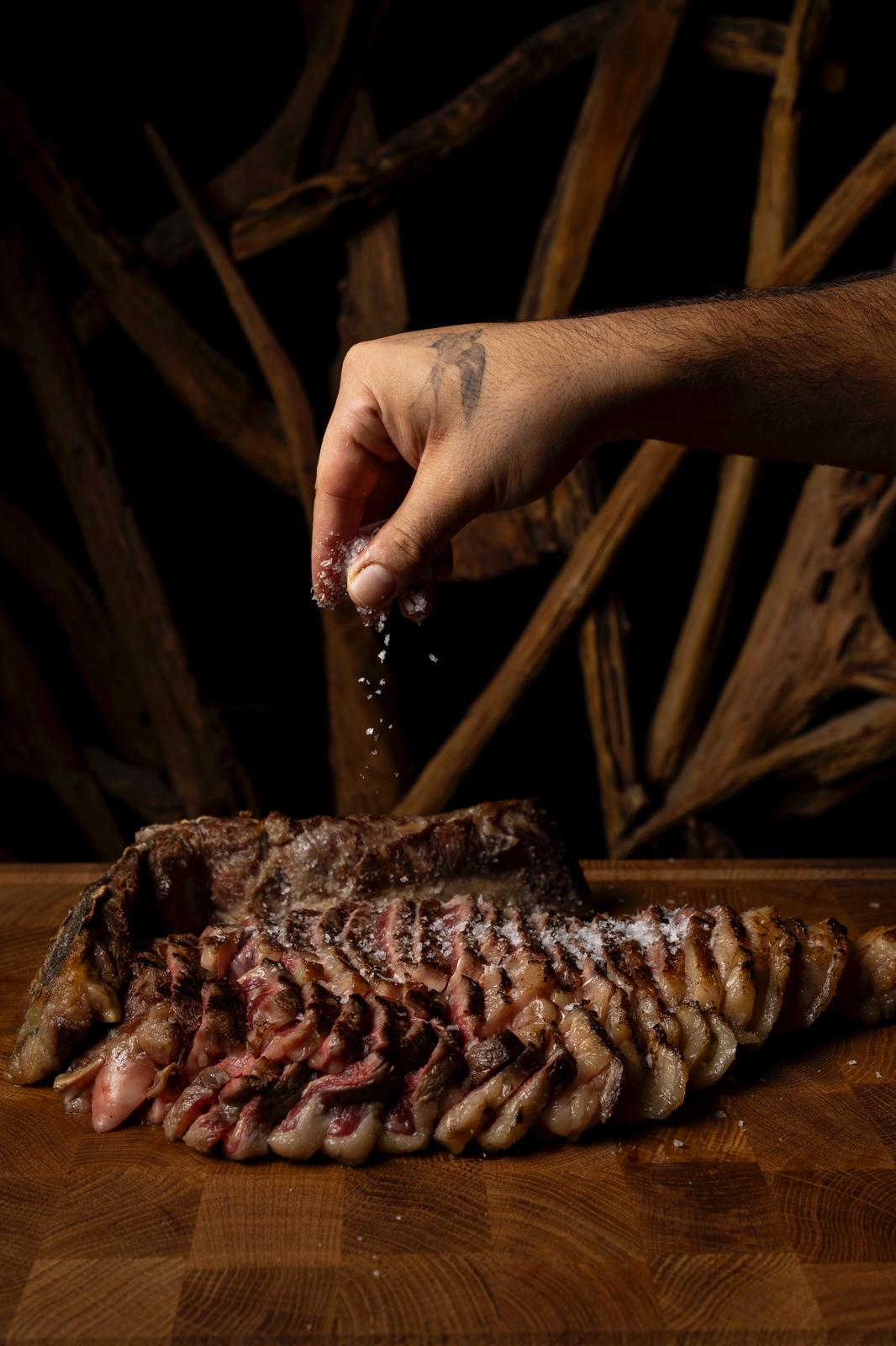 Una mano espolvorea sal sobre carne cortada en una tabla, con ramas secas al fondo